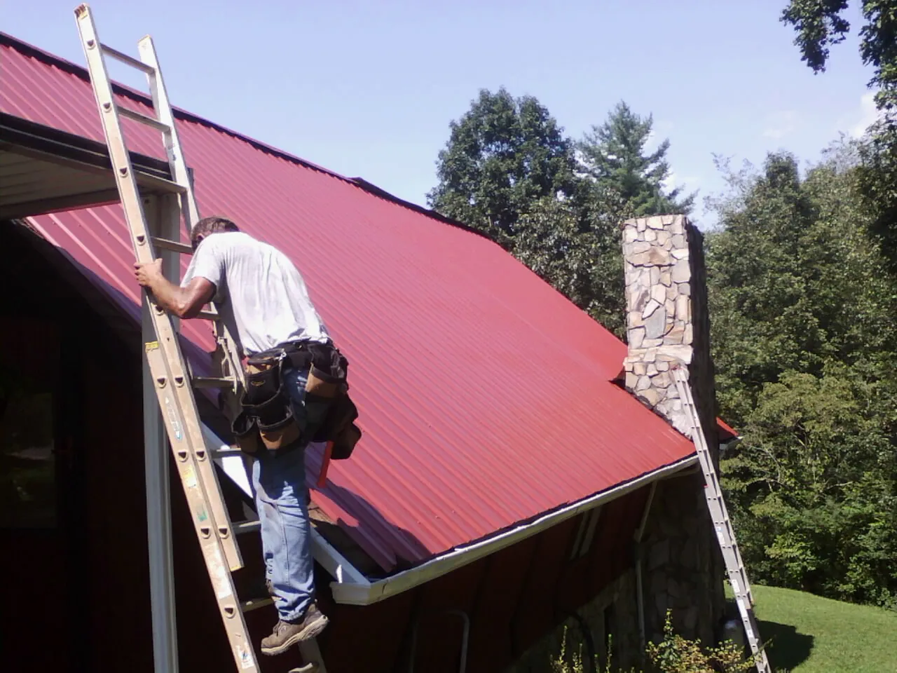 Roofer on a ladder beside a red metal roof and stone chimney — Soffit & Fascia Repair by Roofing Brothers in Allegan